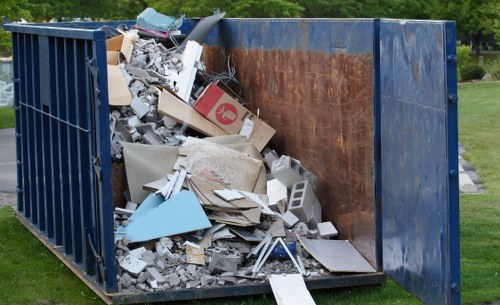 A large, rectangular metal skip with blue sides and a rusted interior, situated outdoors on a grassy area with a background of trees and shrubs. The skip is filled with a mixture of construction and household waste, including broken plasterboard, concrete blocks, cardboard boxes, plastic sheeting, and various discarded materials. The debris is piled high, spilling slightly over the top edges, and some items are leaning or resting against the sides of the skip. The waste appears to be a mix of different textures and colors, predominantly in shades of white, grey, and brown, with some blue and red elements visible on packaging. The surrounding environment suggests a residential or parkland setting, and the overall scene reflects a typical rubbish removal site, consistent with services provided by companies like Commercial Waste Removal Forest Hill.