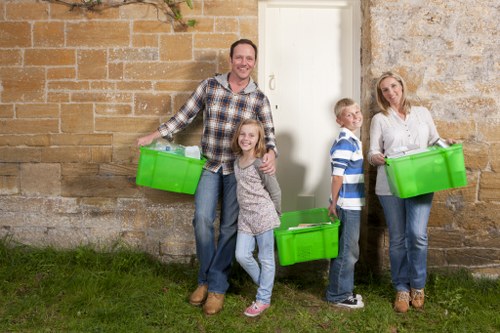 A family of four standing outdoors in front of a stone and brick wall, each holding a bright green plastic container used for rubbish or waste collection. The father, dressed in a checkered shirt and jeans, is smiling and holding a container in his right hand, with a young girl standing in front of him, also smiling, wearing a grey patterned shirt and light jeans. To the right, a boy in a blue and white striped shirt and jeans is holding a container and looking slightly to the side. An adult woman standing further right in a white blouse and blue jeans is also holding a green container and appears to be engaging with the family. The scene takes place on a patch of grass next to the building, which has a mix of stone and brickwork, suggesting a residential or commercial property. The overall atmosphere is casual and positive, aligned with rubbish removal services such as those offered by Commercial Waste Removal Forest Hill, serving local waste management needs in the area.