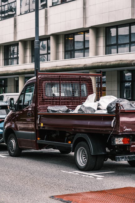 A small burgundy flatbed pickup truck is parked on a city street in front of a modern office building with large glass windows and concrete panels. The truck's cargo bed is filled with various types o