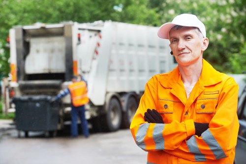 A professional waste management worker in an orange high-visibility jacket and white cap stands with arms crossed in the foreground on a paved driveway, with a confident expression. Behind him, a large white waste collection truck with its rear loading compartment open is visible, with another worker wearing a similar high-visibility safety vest operating nearby and handling a black wheeled bin. The setting is outdoors amidst lush green trees, indicating a suburban or town area, and the surface appears to be a paved driveway or street. The scene depicts rubbish collection or removal activity in a typical local environment, supporting commercial waste removal services, possibly in Forest Hill or surrounding areas. The background emphasizes the environmental context of rubbish collection, with natural daylight illuminating the scene and highlighting the textures of the truck, bin, and safety gear.