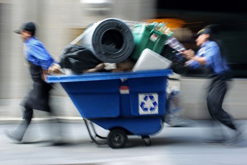 A blue wheeled rubbish bin filled with various waste items, including black garbage bags, a large green plastic container, and other debris, situated outdoors on a paved surface. The bin appears to be in motion, with two individuals dressed in blue uniforms and black caps on either side, pushing it forward. The background is blurred, indicating movement, and the setting may be an urban street or alleyway, possibly in or near Forest Hill. The scene relates to waste collection or rubbish removal services provided by Commercial Waste Removal Forest Hill, highlighting their activities in managing bulky waste and refuse disposal. The waste items on top of the bin include mixed materials and textures such as plastic, metal, and fabric, and the overall environment suggests a service operation in a local residential or commercial area focused on rubbish removal.